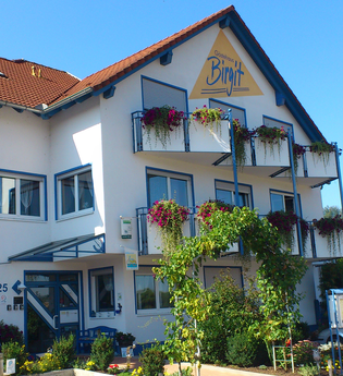 Gästehaus Birgit mit blumengeschmückten Balkonen, grünem Pflanzenbogen am Eingang und mehreren geparkten Autos vor dem Haus | © Gästehaus Birgit