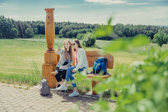 2 Frauen sitzen auf einer Holzbank mit einer Holzsäule und machen ein Selfie. Im Hintergrund sind Wald und Wiesen. Odenwald. | © Tourstikgmeinschaft Odenwald e.V.
