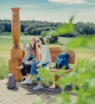 2 Frauen sitzen auf einer Holzbank mit einer Holzsäule und machen ein Selfie. Im Hintergrund sind Wald und Wiesen. Odenwald. | © Tourstikgmeinschaft Odenwald e.V.