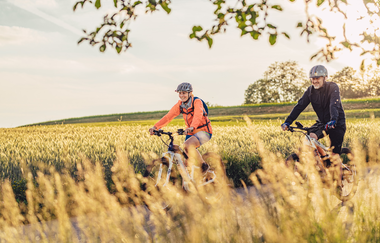 Zwei Radfahrer fahren auf einem Radweg. Im  Hinergrund ist ein Feld mit Getreide zu sehen. Odenwald. | © Touristikgemeinschaft Odenwald e.V.