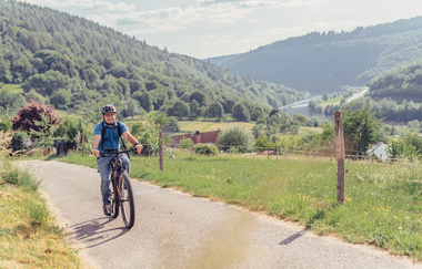 Ein Radfahrer mit blauem Shirt fährt einen Radweg hoch. Im Tald ist ein Fluß mit bergigen und bewaldeten Ufern. Odenwald. | © Touristikgemeinschaft Odenwald e.V.