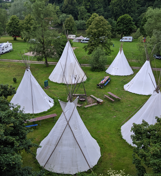 Blick von oben auf eine Wiese mit 6 Tipis die in einem Kreis stehen. In der Mitte ist eine Feuerstelle mit Bänken. Neckargerach im Odenwald. | © ODENWALD river camp