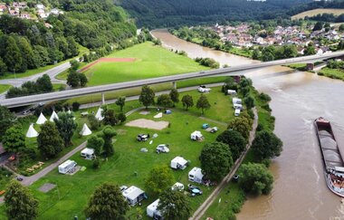 Ein Fluss mit einem Binnenschiff und einer großen Straßenbrücke die über den Fluß geht. Auf der rechten Uferseite ist eine Stadt zu sehen. Auf der linken Seite en Campingplatz mit Zelten und Wohnmobilen. Neckargerach im Odenwald | © ODENWALD river camp