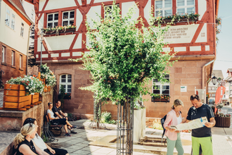 Ein Platz vor einem großen Fachwerkhaus. Auf der rechten Seite steht ein Paar und schaut in eine Karte. Auf der linken Seite stehen zwei Bänke auf welchen Personen sitzen. Walldürn im Odenwald. | © Touristikgemeinschaft Odenwald e.V.