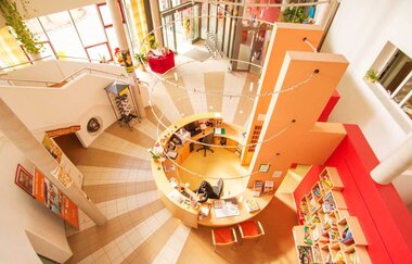 Blick von oben auf einen runden Empfangstresen in einem hellen, modernen Innenraum mit orangefarbenen Wänden und Bücherregalen | © Savoy Hotel