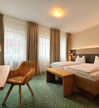 Ein Doppelzimmer im Hotel mit grünem Teppichboden. Ein Tisch mit einem Sessel stehen vor dem Bett. | © Stadthotel Münze