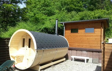 Fass-Sauna mit schwarzem Dach und Holzverkleidung neben einem kleinen Holzhaus (Sauna), umgeben von Bäumen | © Vitalhotel König