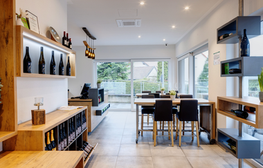 Moderne Vinothek mit Verkaufsraum und Sitzgelegenheiten, dekoriert mit Holzelementen, Weinflaschenleuchte und hauseigenen Weinerzeugnissen im Schaufenster und Ausstellungsraum | © Touristikgemeinschaft Hohenlohe | Heinz J. Schwab