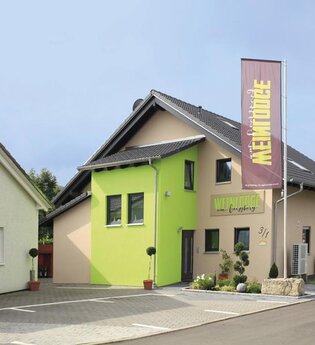 Gebäude von Außen zu sehen mit grünem und braunen Farben und einer großen Fahne im Vordergrund mit Weinlodge