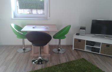 Raum mit kleinem Tisch, drei Stühlen, Fenster, grünem Teppich und TV auf TV Board