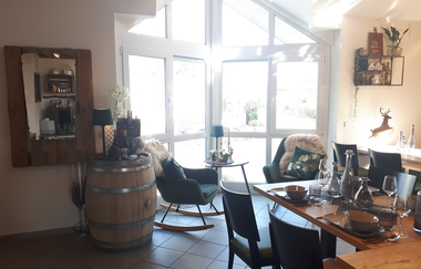 Vinothek mit mehreren Sitzmöglichkeiten und großer Fensterfront im Hintergrund, davor ein Weinfass mit Lampe darauf | © Weinlodge am Geisberg