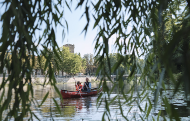 Das Bild wurde zwischen den hängenden Blättern einer Trauerweide aufgenommen. Zu erkennen sind zwei Personen in einem Kanu auf dem Fluss. Im Hintergrund erkennt man einen Turm aus Naturstein | © Heilbronn Marketing GmbH