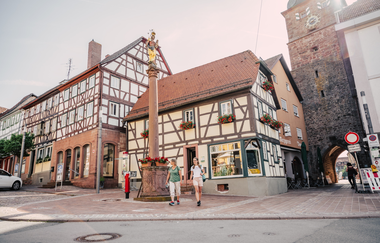 Zwei Fachwerkhäuser mit einer Sandsteinsäule im Vordergrund auf der eine goldene Figur ist. 2 Frauen die an der Säule vorbeilaufen. Neben den Fachwerkhäusern ist ein Stadtturm mmit Uhr. Buchen im Odenwald | © Touristikgemeinschaft Odenwald e.V.