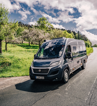 Dunkler Kastenwagen fährt auf einer Straße. Links vom Wohnmobil ist eine Streuobstwiese.| Odenwald | © Touristikgemeinschaft Odenwald e.V.