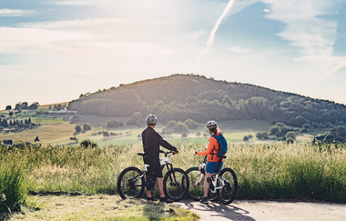Zwei Radfahrer stehen vor einer Wiese und schauen auf eine Erhebung. Waldbrunn im Odenwald. | © Touristikgemeinschaft Odenwald e.V.