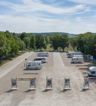 Luftaufnahme vom Stellplatz. Im Vordergrund wird der Platz durch Begrüßungstafeln abgegrenzt, Wohnmobile stehen auf dem Schotterplatz. | © Bad Mergentheim