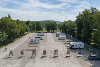 Luftaufnahme vom Stellplatz. Im Vordergrund wird der Platz durch Begrüßungstafeln abgegrenzt, Wohnmobile stehen auf dem Schotterplatz. | © Bad Mergentheim