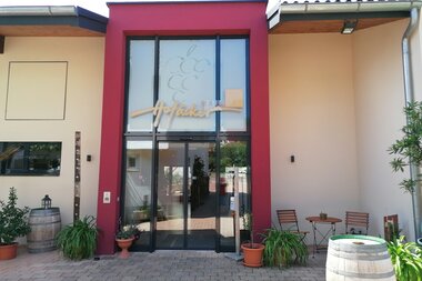 Blick auf den Eingangsbereich mit Glasfront vom Weingut Hofäcker in Weikersheim-Queckbronn. Im Hof davor ist ein Sitzgruppe sowie Blumen und Weinfässer als Dekoration zu sehen. | © Touristinfo Weikersheim
