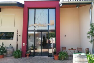 Blick auf den Eingangsbereich mit Glasfront vom Weingut Hofäcker in Weikersheim-Queckbronn. Im Hof davor ist ein Sitzgruppe sowie Blumen und Weinfässer als Dekoration zu sehen. | © Touristinfo Weikersheim