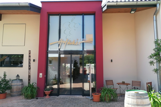 Blick auf den Eingangsbereich mit Glasfront vom Weingut Hofäcker in Weikersheim-Queckbronn. Im Hof davor ist ein Sitzgruppe sowie Blumen und Weinfässer als Dekoration zu sehen. | © Touristinfo Weikersheim