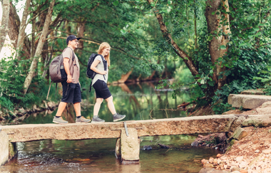 Ein Bach mit einer kleinen Steinbrücke. auf dieser laufen eine Frau und ein Mann. Im Hintergrund sind die Uferseiten stark bewachsen. Elztal im Odenwald | © Touristikgemeinschaft Odenwald e.V.