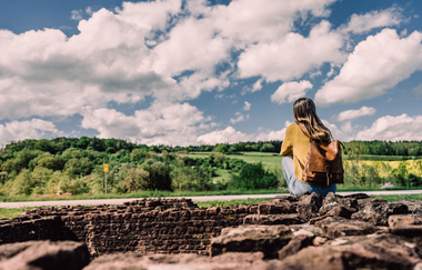 Eine Frau mit gelbem Pullover sitzt auf den Mauern eines Römerkastells und blickt auf eine Landschaft mit Bäumen und Wiesen. Elztal im Odenwald. | © Touristikgemeinschaft Odenwald e.V.