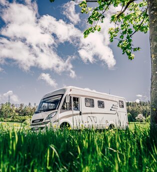 Weißes Wohnmobil fährt auf einer Straße. Im Vordergrund ist eine Blumenwiese im Hintergrund eine Streuobstwiese mit angrenzendem Wald. | Odenwald