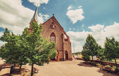 Eine große Kirche aus Buntsandstein mit einem hohen Kirchturm. Ein Platz or der Kirche mit Bäumen. Höpfingen im Odenwald. | © Touristikgemeinschaft Odenwald e.V.