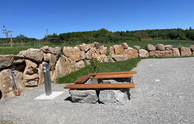 Ein geschotterter Platz mit einer Sitzgelegenheit aus Holz. Die Umgrenzung des Platzes ist aus Findlingsteinen. Im Hintergrund sind Wiesen und Wälder. Limbach im Odenwald. | © Wohnmobilstellplatz Schuler