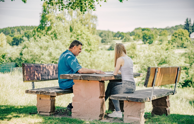 Ein mann und eine Frau sitzen an einem Sitzgelegenheit aus Stein. Im Hintergrund sind Wiesen und Felder. Mudau im Odenwald. | © Touristikgemeinschaft Odenwald e.V.