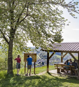 Auf der rechten Seite ist eine Wanderunderstand mit 3 sitzenden Personen. Daneben ist eine Infotafel. Davor stehen ein Mann und eine Frau. Odenwald. | © Touristikgemeinschaft Odenwald e.V.