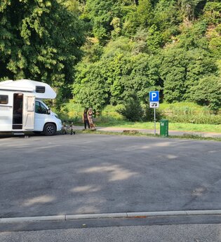 Geteerter Wohnmobilstellplatz mit einem weißen Wohnmobil daneben stehen zwei Frauen. Hinter dem Wohnmobil fliesst der Neckar. | Neckargerach | Odenwald | © Gemeinde Neckargerach