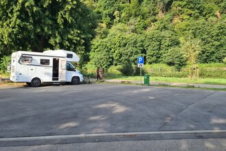 Geteerter Wohnmobilstellplatz mit einem weißen Wohnmobil daneben stehen zwei Frauen. Hinter dem Wohnmobil fliesst der Neckar. | Neckargerach | Odenwald | © Gemeinde Neckargerach