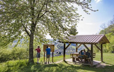 Auf der rechten Seite ist eine Wanderunderstand mit 3 sitzenden Personen. Daneben ist eine Infotafel. Davor stehen ein Mann und eine Frau. Odenwald. | © Touristikgemeinschaft Odenwald e.V.