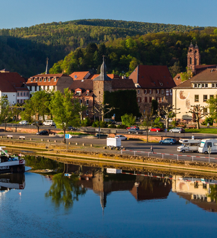 Im Vordergrund ist ein Fluß mit einem Binnenschiff. Dahinter am Ufer ist direkt ein Wohnmobilstellplatz mit 3 Wohnmobilen. Im Hintergrund ist eine Stadt mit Häusern und einer Kirche. Eberbach im Odenwald. | © Stadtverwaltung Eberbach