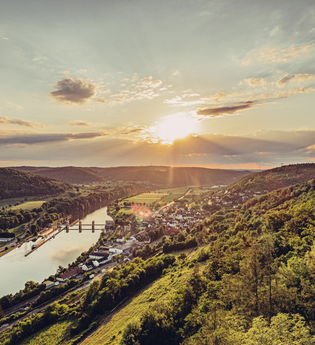 Weitblick mit Sonnenuntergang auf einen Fluss. Auf der rechten Uferseite erstreckt sich eine Gemeinde. Neckarzimmern im Odenwald. | © Touristikgemeinachaft Odenwald e.V.