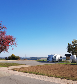 Blick auf einen Parkplatz mit einem Wohnmobil. Daneben steht eine Infotafel, ein Baum und eine Sitzgelegenheit. Neunkirchen im Odenwald.