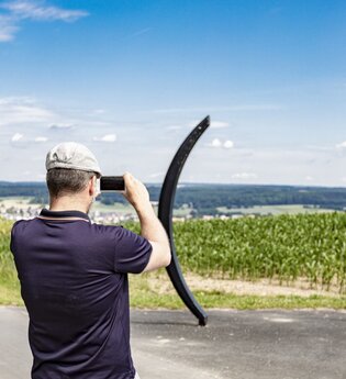 Ein Mann mit Fernglas schaut in die Ferne. Vor ihm stehen zwei halbrunde Gebilde, die aussiehen wie eine große Klammer. Dahinter ist ein grünes Feld. Osterburken im Odenwald. | © Touristikgemeinschaft Odenwald e.V.