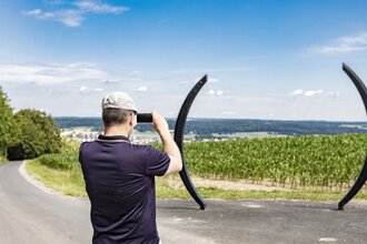 Ein Mann mit Fernglas schaut in die Ferne. Vor ihm stehen zwei halbrunde Gebilde, die aussiehen wie eine große Klammer. Dahinter ist ein grünes Feld. Osterburken im Odenwald. | © Touristikgemeinschaft Odenwald e.V.