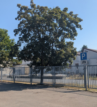 Es ist ein Parkplatz mit einem Wohnmobilschild zu sehen, begrenzt durch einen Zaun. Hinter dem Zaun steht ein Fabrikgebäude. Odenwald. | © Gemeinde Schefflenz