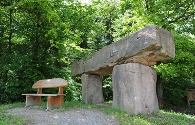 Man sieht einen kleinen Platz im Wald mit einer Holzbank. Daneben stehen 3 große Findlinge die wie eine überdimensionale Brücke angeordnet sind. Schönbrunn im Odenwald | © Touristikgemeinschaft Odenwald e.V.