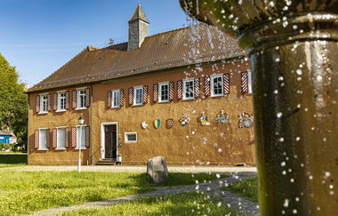 Man sieht ein Schloss mit kleinem Türmchen und Holzläden an den Fenstern. Im Vordergrund sieht man ein Teil eines Brunnen mit perlendem Wasser. Odenwald. | © Touristikgemeinschaft Odenwald e.V.