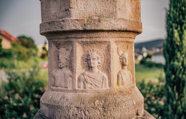 Ein Ausschnitt einer Buntsandsteinsäule mit römischen Männern. Im Hintergrund eine verschwommene Stadt mit Wäldern. Odenwald. | © Touristikgemeinschaft Odenwald e.V.