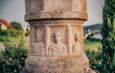 Ein Ausschnitt einer Buntsandsteinsäule mit römischen Männern. Im Hintergrund eine verschwommene Stadt mit Wäldern. Odenwald. | © Touristikgemeinschaft Odenwald e.V.