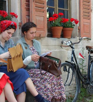 Drei Frauen sitzen vor einem Haus. Sie tragen Kleidung, die ins Jahr 1947 passt. Neben den Frauen lehnt ein Fahrrad an die Hauswand. Die Frauen haben alle je eine Tasche auf dem Schoss. Sie holen etwas aus ihren Handtaschen raus. | © Hohenloher Freilandmuseum