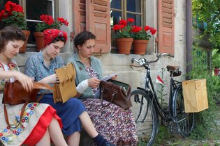 Drei Frauen sitzen vor einem Haus. Sie tragen Kleidung, die ins Jahr 1947 passt. Neben den Frauen lehnt ein Fahrrad an die Hauswand. Die Frauen haben alle je eine Tasche auf dem Schoss. Sie holen etwas aus ihren Handtaschen raus. | © Hohenloher Freilandmuseum