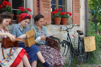 Drei Frauen sitzen vor einem Haus. Sie tragen Kleidung, die ins Jahr 1947 passt. Neben den Frauen lehnt ein Fahrrad an die Hauswand. Die Frauen haben alle je eine Tasche auf dem Schoss. Sie holen etwas aus ihren Handtaschen raus. | © Hohenloher Freilandmuseum