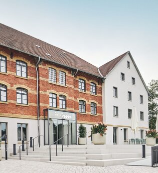 Zwei Gebäude, eines aus rotem Backstein und eines weiß, mit einem Eingang und Treppen. Blumenkübel und Tische mit Sonnenschirmen sind vorhanden. | © Kocherwerk