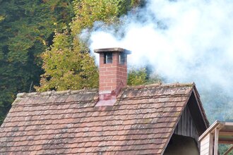 Aus einem Schornstein auf einem  Dach steigt Rauch. Der Rauch ist weiß. Das Dach ist mit roten Ziegeln gedeckt. Es ist Teil eines historischen Backofens.