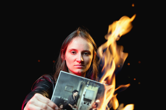 Eine junge Frau hält ein brennendes Foto in die Kamera. Auf dem Bild sieht man eine Familie am Küchentisch sitzen, die Mutter bedient den Vater.